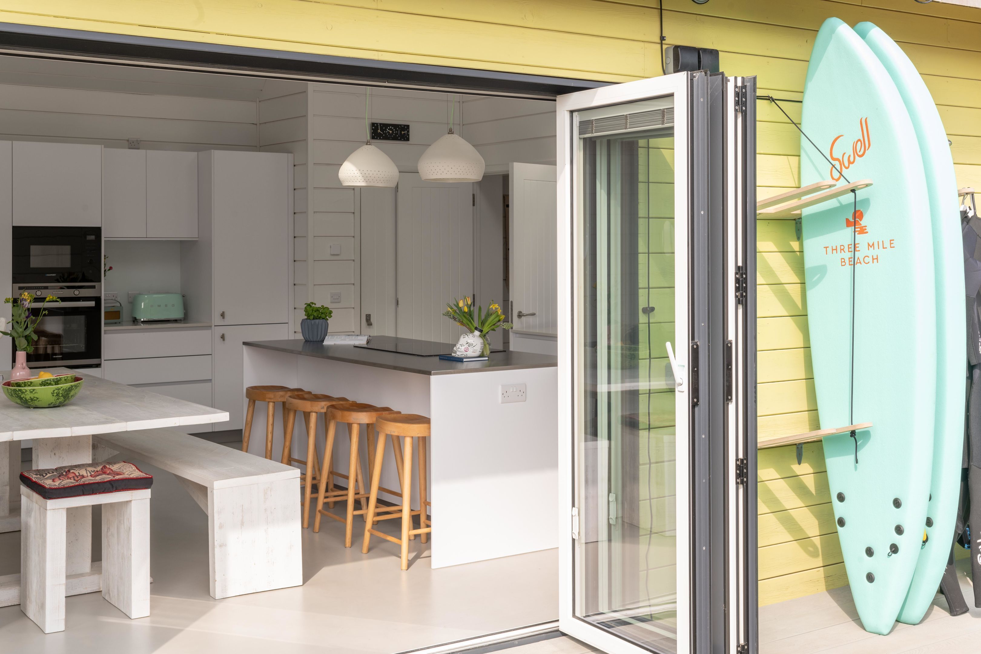 A modern kitchen with white cabinets, a central island with stools, pendant lighting, and a surfboard on the wall.