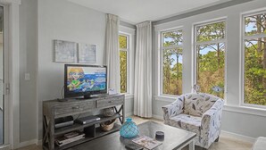 Ein Wohnzimmer mit Fernseher, Couchtisch, einem gemusterten Sessel und großen Fenstern mit Blick auf Bäume.