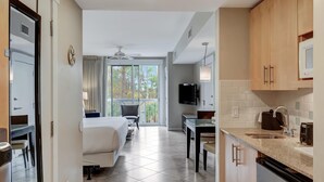 Une chambre d’hôtel comprenant un lit, un bureau, une chaise, une télévision, une fenêtre avec des rideaux et un coin cuisine avec un évier et des placards.
