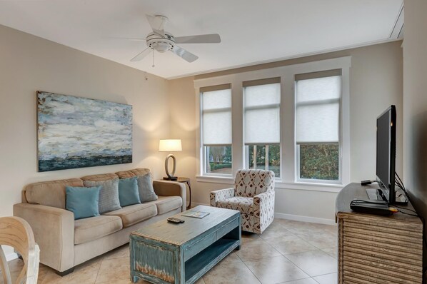 A living room with a ceiling fan, a large painting, a sofa with cushions, a patterned armchair, a coffee table, a lamp, and a TV on a wooden cabinet.