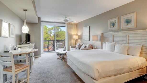 A hotel room with a large bed, a desk with a chair, a small table, a ceiling fan, and a balcony with a view of trees.