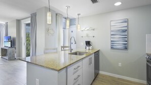 A modern kitchen with a granite countertop, stainless steel sink, and a built-in dishwasher. There are pendant lights, a coffee maker, and a framed artwork on the wall.