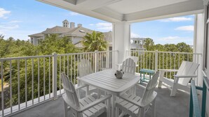 A covered balcony with white furniture, including a table and chairs, overlooking a residential area with houses and trees.