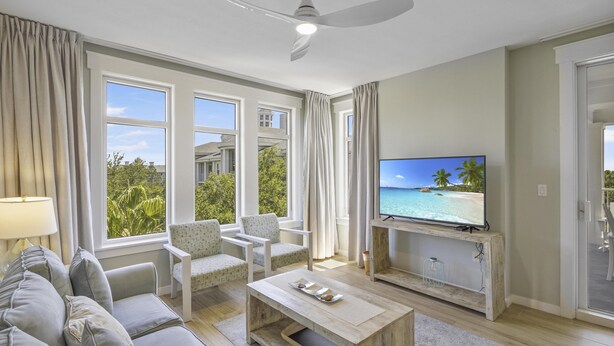 A living room with a sofa, chairs, a coffee table, a TV, and large windows with curtains.