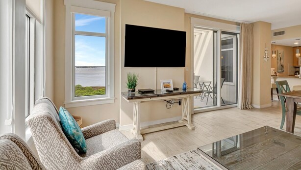Un vaste salon doté d’un grand téléviseur à écran plat, d’un coin repas avec une table et des chaises, et offrant une vue sur un plan d’eau grâce à de grandes fenêtres.
