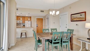 Un coin repas avec une table en bois et des chaises vertes, un lustre et une cuisine en arrière-plan.