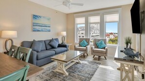 Un salon comprenant un canapé, des fauteuils, une table basse et un téléviseur. Il y a des fenêtres donnant sur des immeubles.