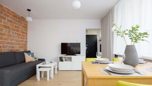 Una sala moderna con pared de ladrillo, televisor de pantalla plana, zona de comedor con mesa para dos comensales y un sofá con cojines.