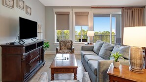 A living room with a flat-screen TV, a wooden cabinet, a patterned armchair, a grey sofa, a coffee table, and a balcony with a view of trees.