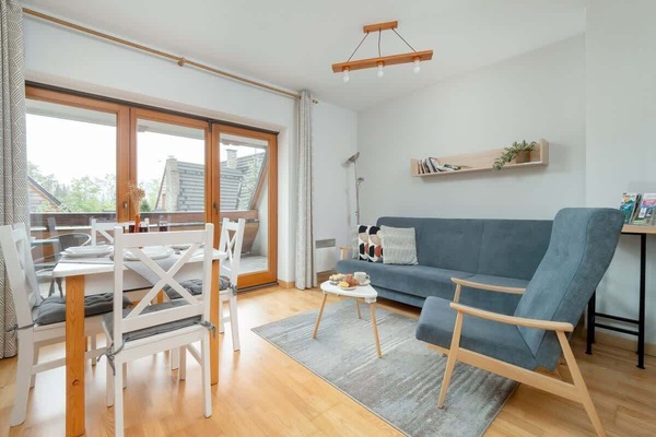 A bright living room with wooden floors, a blue sofa, a white dining table, and large balcony doors.