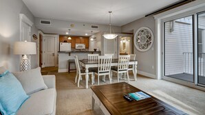 Amplia sala de estar con sofá, mesa de centro y comedor con mesa y sillas. Se observa una cocina al fondo con armarios y electrodomésticos.