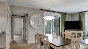 Un comedor con mesa de madera y sillas blancas, un espejo decorativo grande y un televisor sobre un mueble.