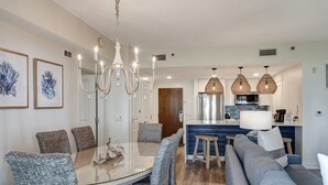 A modern kitchen with a central island, a dining area with a glass table and wicker chairs, and a living area with a gray sofa and blue cushions.