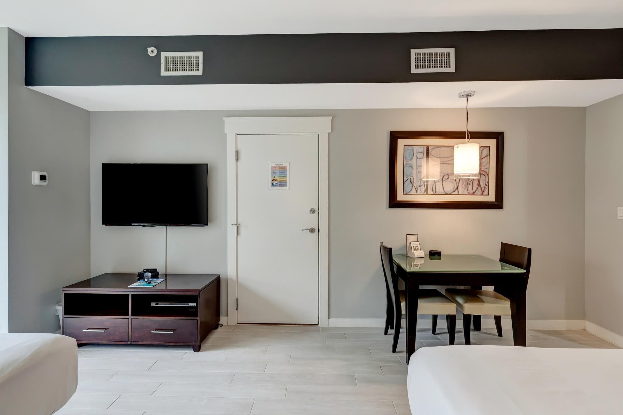 Habitación de hotel con televisor de pantalla plana, una mesa de comedor con sillas, una cama y una obra de arte enmarcada sobre la mesa.