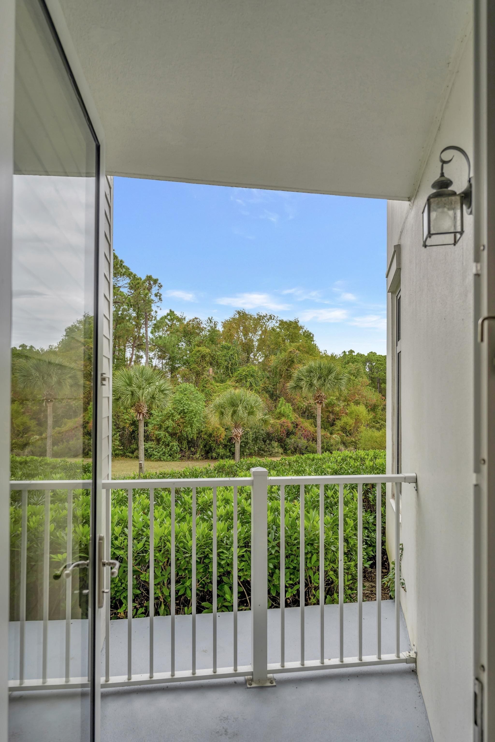A balcony with a white railing, a glass door, and a view of a lush garden with various plants and trees.