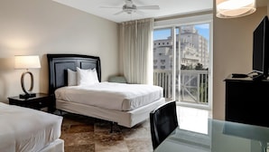 Habitació d'hotel amb llit, escriptori, cadira, televisió, ventilador de sostre i finestra amb vista a edificis.