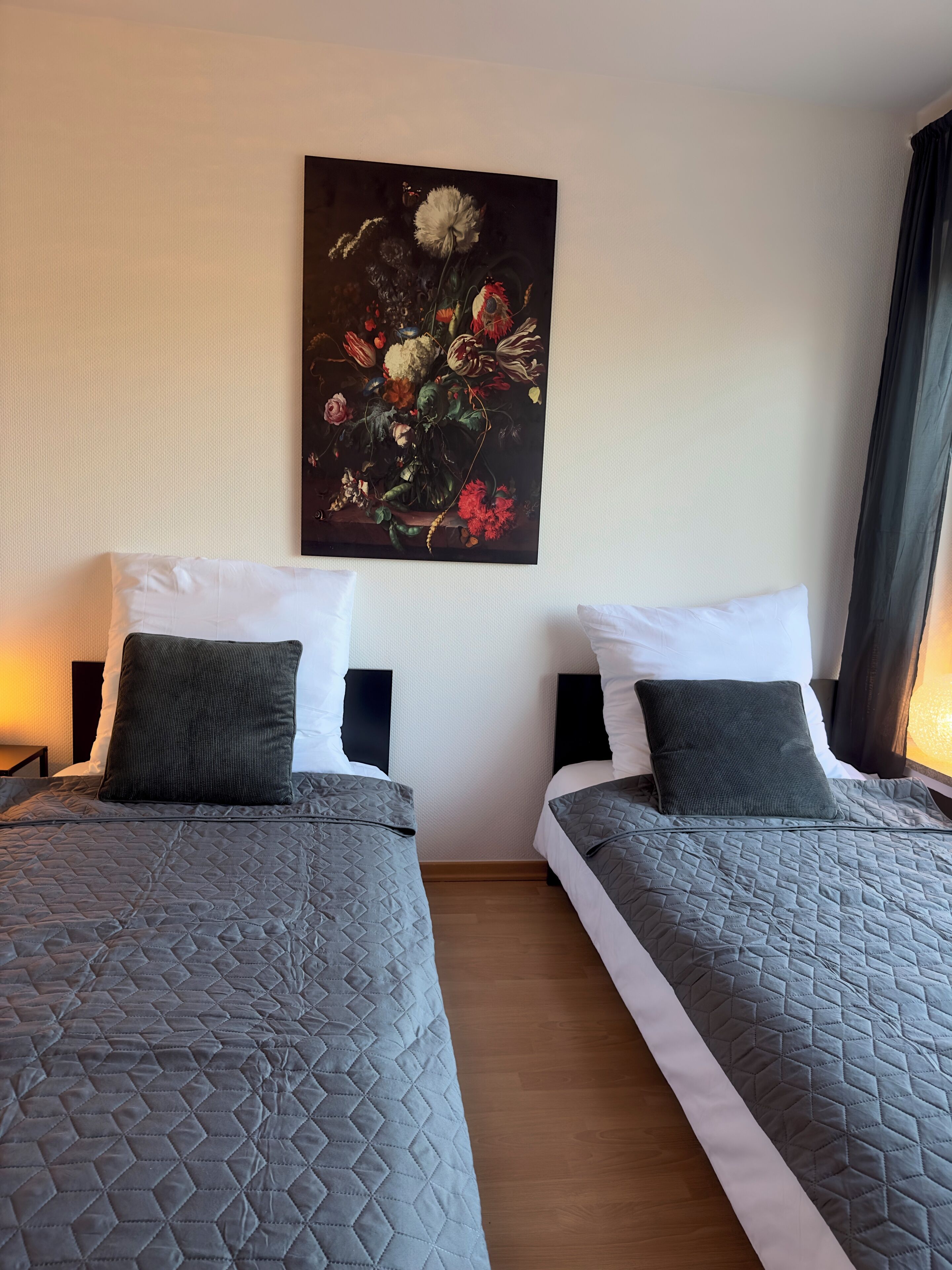 Une chambre d’hôtel avec deux lits, chacun recouvert d’une courtepointe grise matelassée et agrémenté d’oreillers vert foncé. Un grand tableau représentant des motifs floraux est accroché au mur, au-dessus des lits.