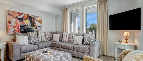 A modern living room with a sectional sofa, ottoman, and floral armchair. A ceiling fan, large windows with curtains, and a flat-screen TV are visible.