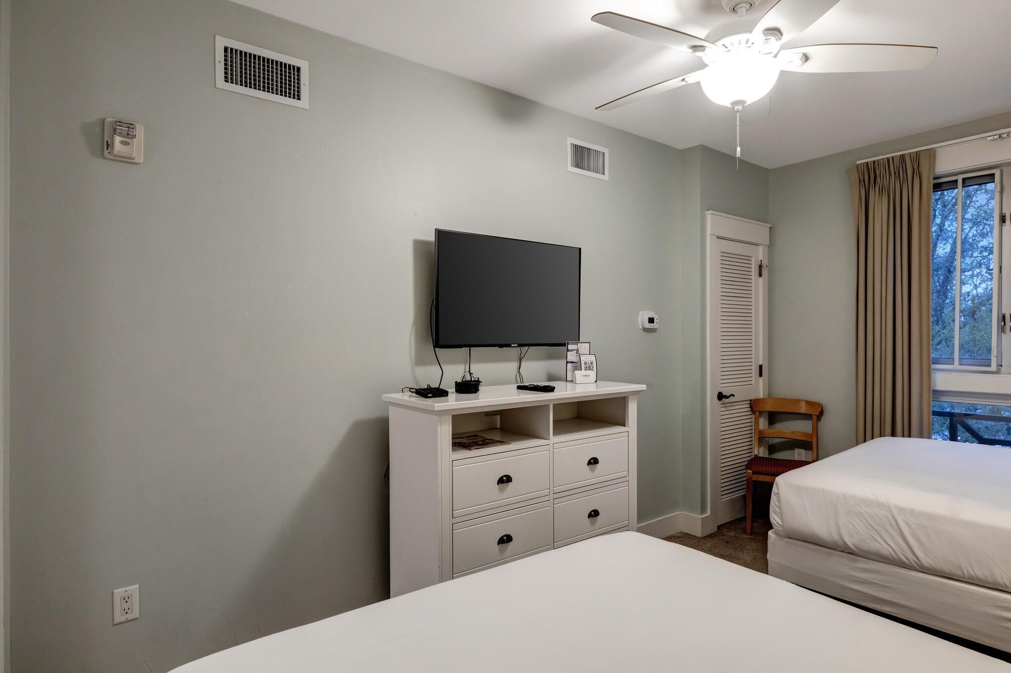 Un dormitorio con una cama, un televisor sobre un tocador blanco, un ventilador de techo y una ventana con cortinas.