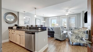 A modern kitchen with a central island, wooden cabinets, and a dishwasher. There is a seating area with a sofa and chairs, and large windows providing natural light.