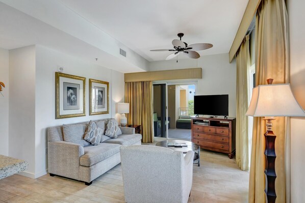 A living room with a sofa, armchair, ceiling fan, television, and a view of the outside.