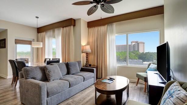 A hotel room with a gray sofa, a wooden coffee table, a TV, and a large window with a view of buildings. - Relaxing Oasis in Luau: Serene Two-Bedroom Retreat with 3 Beds and Scenic Views at Sandestin Golf and Beach Resort! (Miramar Beach)