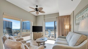 Amplia sala de estar con un televisor de pantalla plana grande, asientos cómodos, ventilador de techo y vistas de la ciudad a través de amplios ventanales.