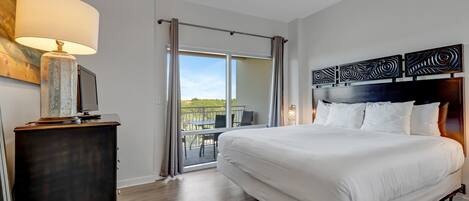 Habitación de hotel con una cama grande, un escritorio con lámpara, televisor y un balcón con mesa y sillas.