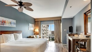 A hotel room with a large bed, a desk with a chair, a ceiling fan, and a view of buildings through the window.
