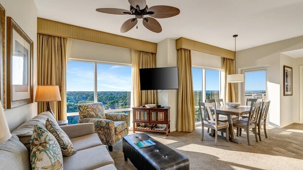 Ein geräumiges Wohnzimmer mit großem Flachbildfernseher, Deckenventilator, einer Couch mit gemusterten Kissen, einem Esstisch mit Stühlen und großen Fenstern mit Blick auf die Landschaft.
