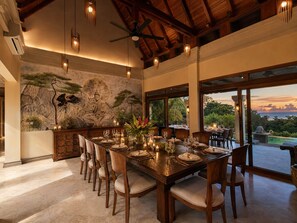 A dining area with a large table set for a meal, surrounded by chairs, with a view of the outdoors. - 5-Acre Private Jungle Estate | Ocean Views, Huge Pool, Chef & Transportation (Playa Tamarindo)