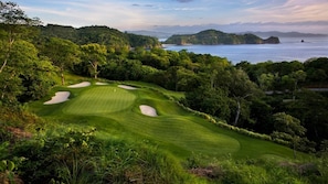 A golf course with sand bunkers, surrounded by dense greenery and overlooking a body of water.