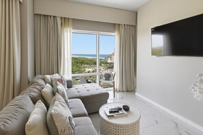 A modern hotel room with a large window, a sofa, a round coffee table, and a flat-screen TV.