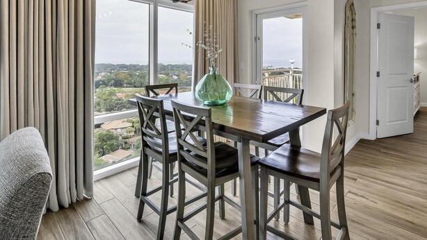 Comedor con mesa de madera, sillas y un jarrón. Amplias ventanas ofrecen vistas a la ciudad.
