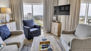 Una sala de estar moderna con sofá, sillón y mesa de centro. Un televisor de pantalla plana está montado en la pared, y una ventana ofrece vistas a la ciudad.