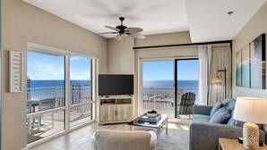 A spacious living room with a large TV, comfortable seating, and a balcony with a view of the ocean.