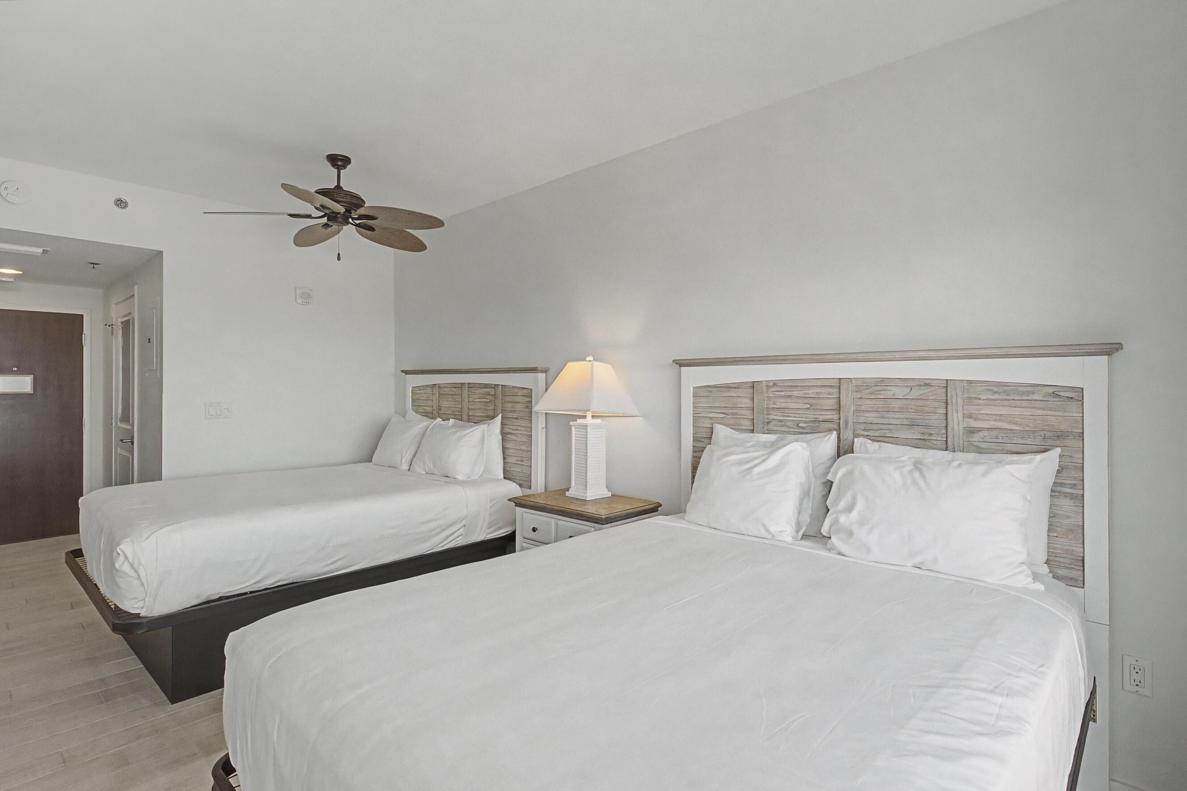 Habitación de hotel con dos camas, ventilador de techo, lámpara de noche y cabecero de madera.