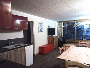 Uma cozinha compacta com lava-louças, uma mesa de jantar com cadeiras, um banquinho vermelho, uma TV sobre um armário de madeira e uma janela com vista para árvores e montanhas.