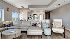 Un salon moderne avec un canapé d’angle, un pouf et une table basse ronde. On aperçoit un coin repas avec une chaise blanche et une petite table d’appoint. La cuisine, avec ses meubles blancs et ses appareils en acier inoxydable, se trouve au fond de la pièce.