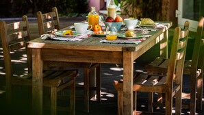 A wooden table set with breakfast items, surrounded by chairs, in a shaded outdoor area with greenery and a building in the background. - Quinta do Ameal (Ponte de Lima)