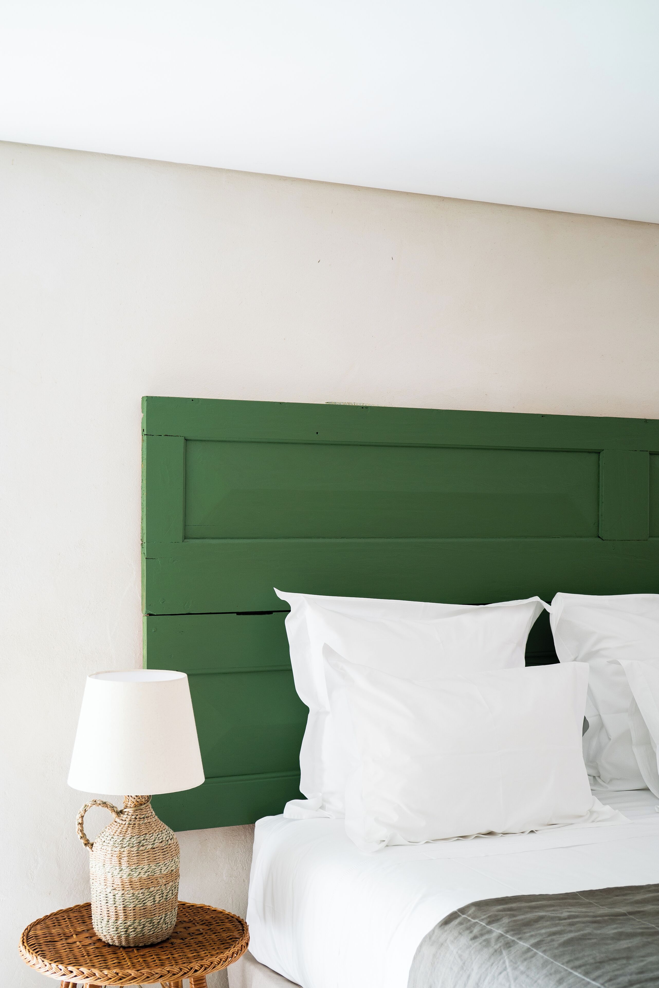 A bed with a green headboard, white bedding, and a woven lamp on a round table beside the bed.