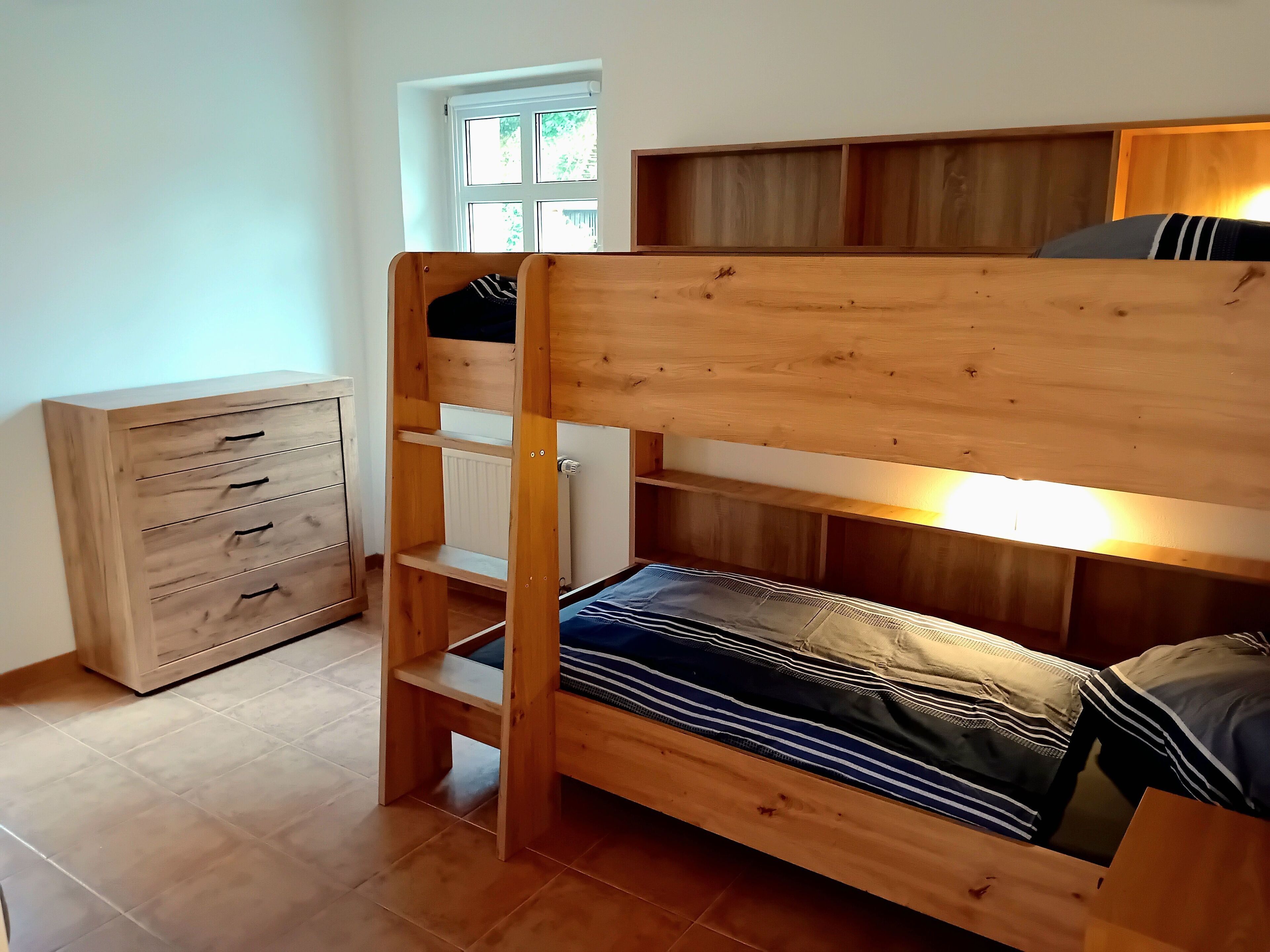Ein Stockbett mit Holzrahmen, eine Kommode mit Schubladen und ein Fenster mit Blick auf Bäume.