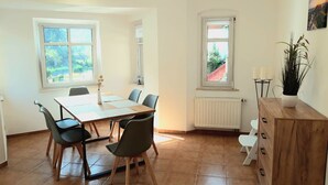 A dining area with a wooden table and chairs, a radiator, and a window with a view of greenery.