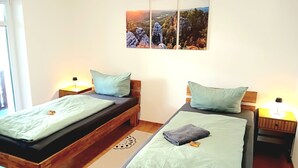 A room with two wooden beds, each with a green mattress and matching pillows. There is a wall-mounted artwork depicting a landscape scene.