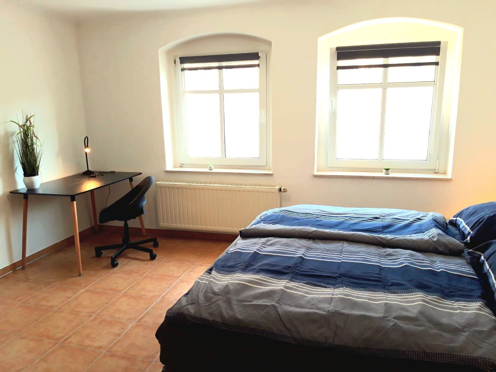 Ein Schlafzimmer mit einem Bett, einem Schreibtisch und zwei Fenstern.