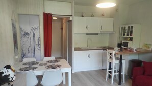Cocina compacta con armarios blancos, microondas y fregadero. Hay una zona de comedor con una mesa para dos, sillas y una cortina roja.