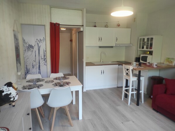 Cocina compacta con armarios blancos, microondas y fregadero. Hay una zona de comedor con una mesa para dos, sillas y una cortina roja.