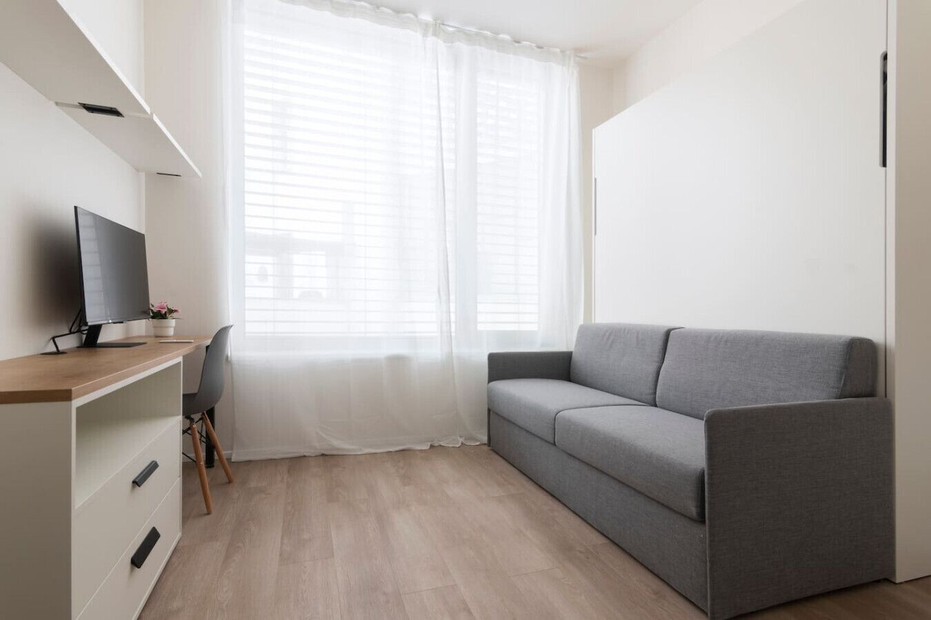 A modern room with a grey sofa, a wooden desk with a computer, and a window with sheer curtains.
