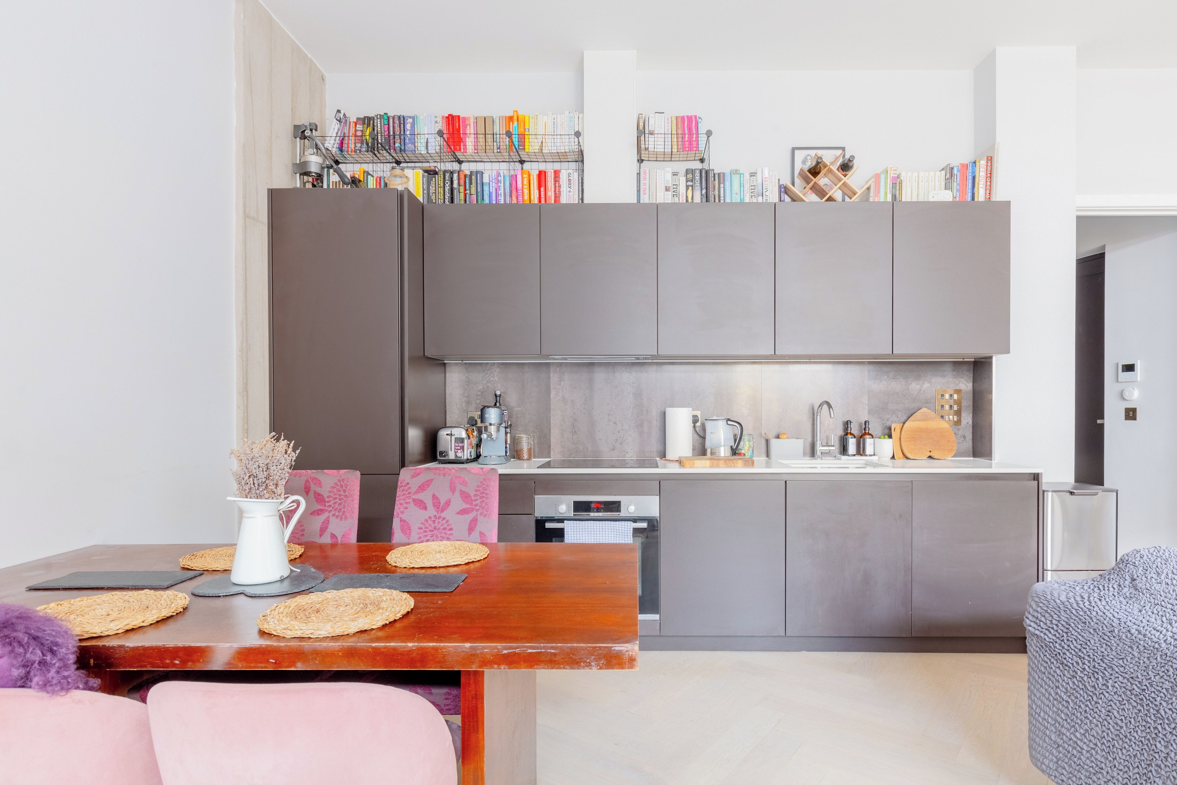 A modern kitchen with grey cabinets, a dining table with a vase and placemats, and a bookshelf above the counter.