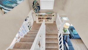 A staircase leading to a living area with a bookshelf, a surfboard, and a painting of a wave. - AMI's Salty Breeze: 5-min walk to Beach, community pool, relaxing decor (Holmes Beach)